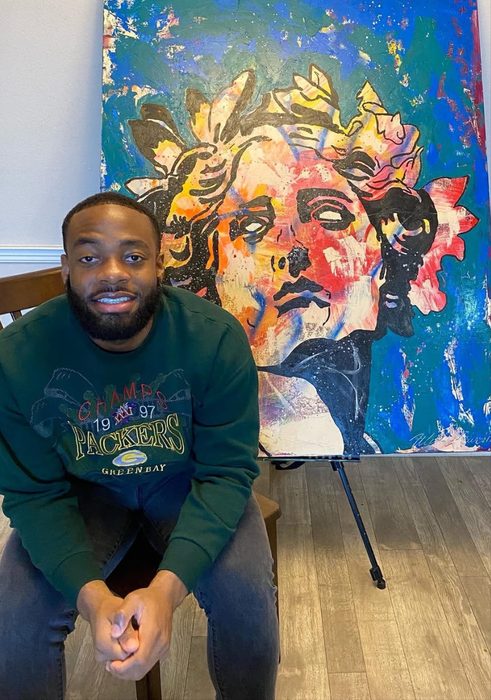 Willie Austin seated in front of his painting of a crowned classical bust in blue, orange and red
