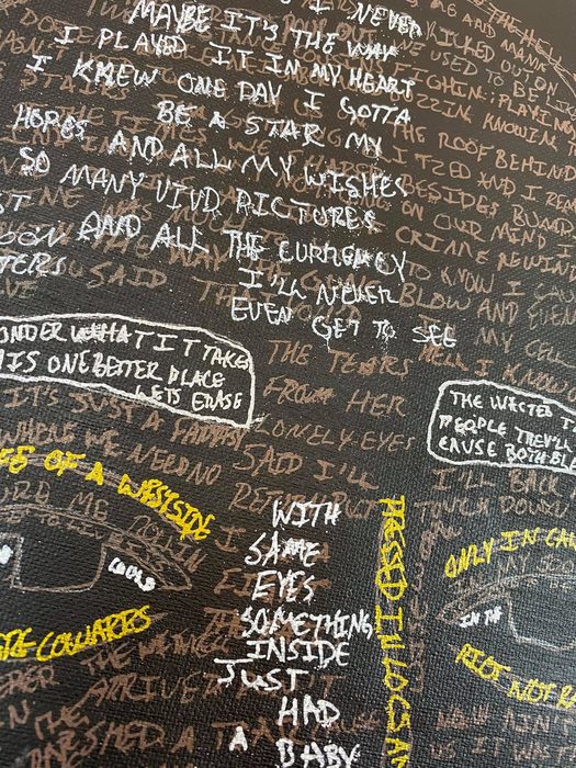 Close view of the canvas, where lines of handwritten lyrics in white, brown and yellow form the face