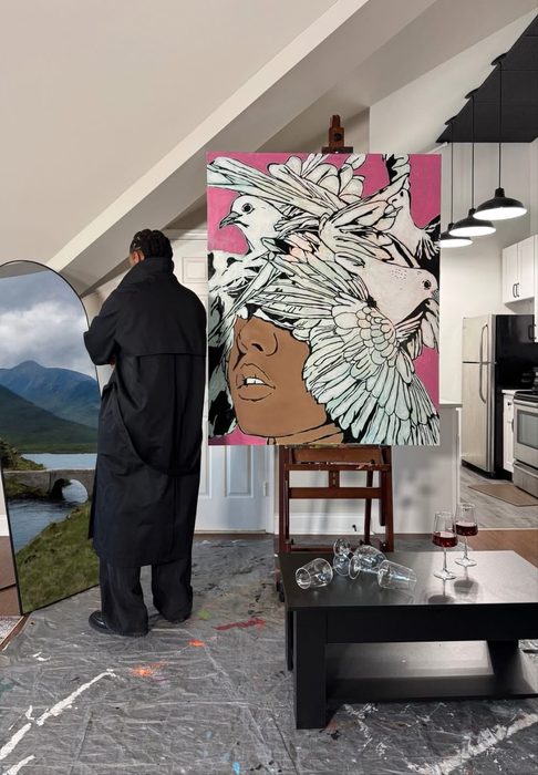 Willie Austin in a long black coat beside his painting of a face beneath a flock of white doves on pink