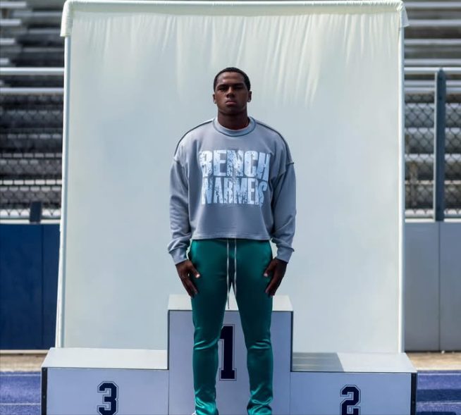 A model on a first place podium in a gray Bench Warmers sweatshirt and green pants