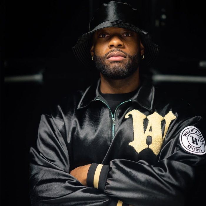 Willie Austin, arms crossed, in a black satin Willie Austin Sideline Jacket with a cream W and a black bucket hat